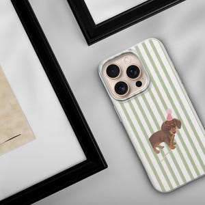 May include: A phone case with a green and white striped pattern. The case features a brown dachshund wearing a pink party hat and holding a party horn. The phone case is on a white surface.