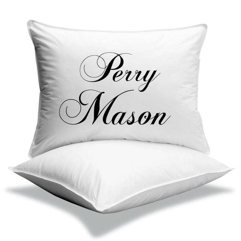 Pode incluir: Duas almofadas quadradas brancas empilhadas uma sobre a outra. A almofada superior tem as palavras "Perry Mason" impressas em elegante escrita preta. As almofadas s&atilde;o feitas de um material macio e liso.