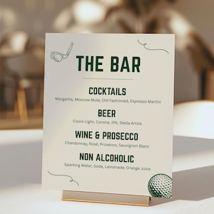 May include: A white bar menu sign with green text and golf-themed illustrations. The sign lists cocktails, beer, wine, prosecco, and non-alcoholic beverages. The sign is held in a gold-colored metal stand.