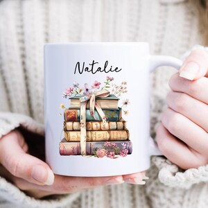 Puede incluir: Taza de cer&aacute;mica blanca con un dise&ntilde;o de libros apilados, flores y una cinta. El nombre "Natalie" est&aacute; escrito en la taza. La taza est&aacute; sujeta por una persona.