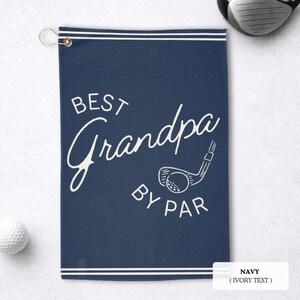 Navy blue golf towel with the text "BEST Grandpa BY PAR" in white script and a golf club graphic. The towel has a white border and a gold-coloured grommet for hanging. A golf ball and club are visible.