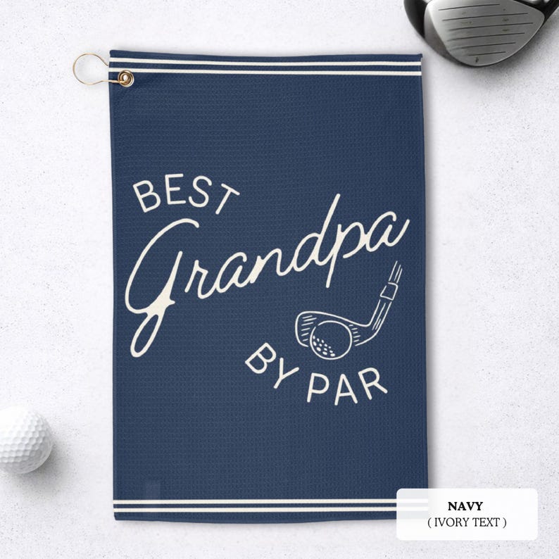 Navy blue golf towel with the text "BEST Grandpa BY PAR" in white script and a golf club graphic. The towel has a white border and a gold-coloured grommet for hanging. A golf ball and club are visible.