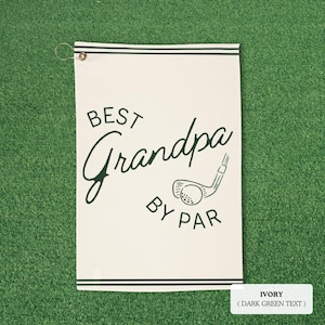 Ivory golf towel with dark green text reading "BEST Grandpa BY PAR" and a golf club and ball graphic. The towel has dark green stripes and a metal grommet for hanging. Measures approximately 40cm x 60cm.