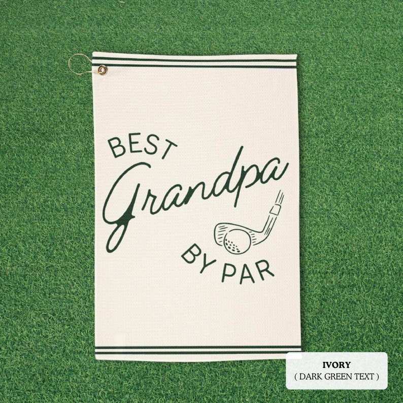 Ivory golf towel with dark green text reading "BEST Grandpa BY PAR" and a golf club and ball graphic. The towel has dark green stripes and a metal grommet for hanging. Measures approximately 40cm x 60cm.