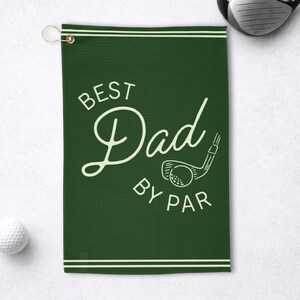 May include: A dark green golf towel with the words "BEST Dad BY PAR" in a cream-colored script. The towel has a golf club and ball graphic and a metal grommet for hanging. A golf ball and club are visible.