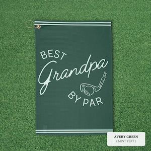 Avery green golf towel with white text reading "BEST Grandpa BY PAR" and a golf club graphic. The towel has a white border and a metal ring for hanging. The background is green.