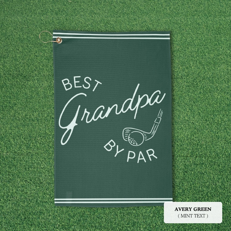 Avery green golf towel with white text reading "BEST Grandpa BY PAR" and a golf club graphic. The towel has a white border and a metal ring for hanging. The background is green.