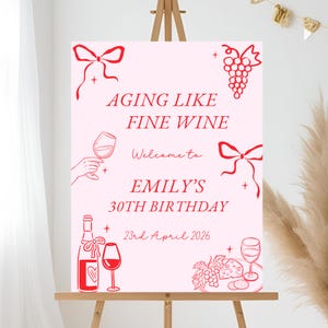 Puede incluir: Un letrero rosa sobre un caballete de madera con ilustraciones y texto en rojo. El letrero dice "AGING LIKE FINE WINE" y da la bienvenida a los invitados a "EMILY'S 30TH BIRTHDAY" el 23 de abril de 2026. Gráficos temáticos de vino decoran el letrero.