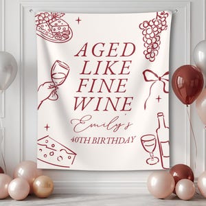 May include: A white tapestry with burgundy illustrations of wine glasses, grapes, cheese, and a wine bottle. Text reads "AGED LIKE FINE WINE Emily's 40TH BIRTHDAY". Balloons in shades of pink and burgundy are in the foreground.