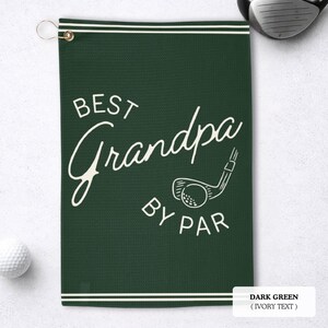 Dark green golf towel with ivory text reading "BEST Grandpa BY PAR". A golf club and ball graphic is also featured. The towel has a hanging loop and white stripes.