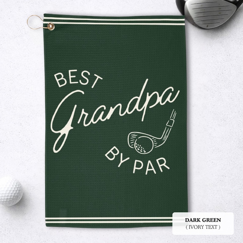 Dark green golf towel with ivory text reading "BEST Grandpa BY PAR". A golf club and ball graphic is also featured. The towel has a hanging loop and white stripes.