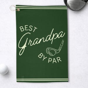 May include: A dark green golf towel with the words "BEST Grandpa BY PAR" in a white script font. A golf club and ball graphic is also featured. The towel has a loop for hanging and white stripes at the top and bottom.