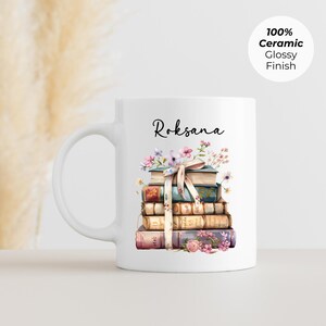 Puede incluir: Taza de cer&aacute;mica blanca con una pila de libros adornada con flores y una cinta. El nombre "Roksana" est&aacute; escrito en cursiva encima de los libros. La taza tiene un acabado brillante y el texto "100% Ceramic Glossy Finish".