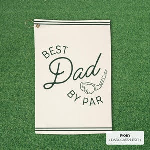 May include: Ivory golf towel with dark green text that reads "BEST Dad BY PAR" with a golf club and ball graphic. The towel has dark green stripes at the top and bottom and a metal grommet for hanging.