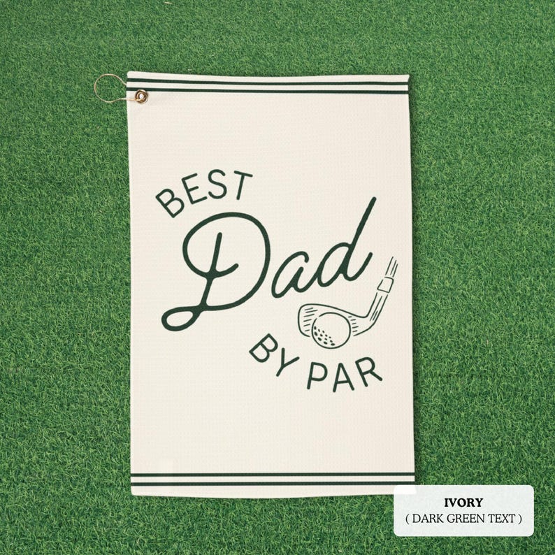 May include: Ivory golf towel with dark green text that reads "BEST Dad BY PAR" with a golf club and ball graphic. The towel has dark green stripes at the top and bottom and a metal grommet for hanging.