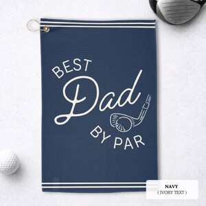May include: Navy blue golf towel with the text "BEST Dad BY PAR" in ivory. The towel features a golf club and ball graphic. A golf ball and club are visible in the background.