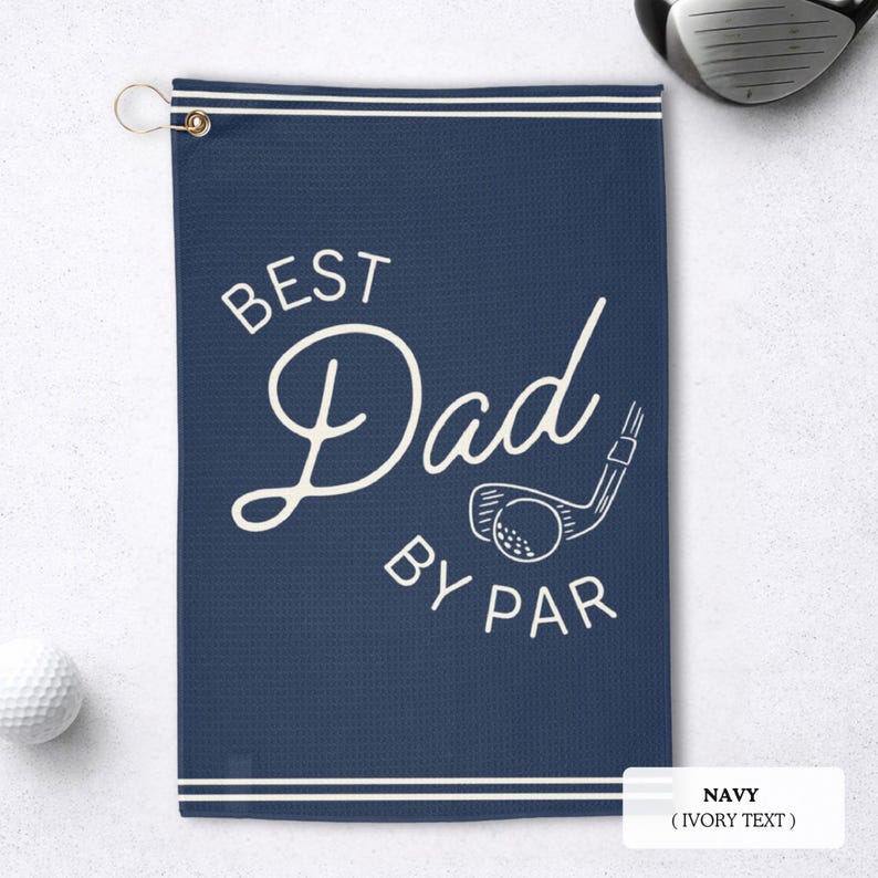 May include: Navy blue golf towel with the text "BEST Dad BY PAR" in ivory. The towel features a golf club and ball graphic. A golf ball and club are visible in the background.