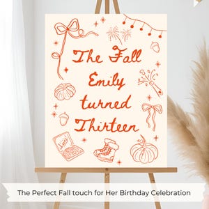 Puede incluir: Un letrero crema sobre un caballete con texto e ilustraciones en naranja. El texto dice "The Fall Emily turned Thirteen". Las ilustraciones incluyen calabazas, lazos y fuegos artificiales. El texto inferior dice "The Perfect Fall touch for Her Birthday Celebration."