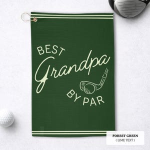A forest green golf towel with the text "BEST Grandpa BY PAR" in a white script font. It features a golf club and ball graphic. The towel has a gold grommet for hanging and white stripes along the top and bottom.