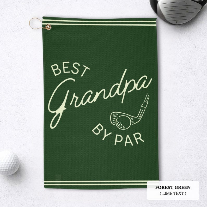 A forest green golf towel with the text "BEST Grandpa BY PAR" in a white script font. It features a golf club and ball graphic. The towel has a gold grommet for hanging and white stripes along the top and bottom.