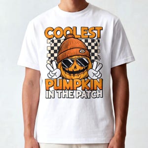 May include: A white t-shirt featuring a graphic of a jack-o'-lantern wearing an orange beanie and sunglasses, making peace signs with white gloved hands. The text reads COOLEST PUMPKIN IN THE PATCH against a black and white checkered background.