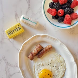May include: A breakfast scene featuring a fried egg, two sausages, and a bowl of raspberries and blackberries. Salt and pepper shakers are visible, along with a butter dish. The egg yolk is bright yellow, and the food is on a white plate.
