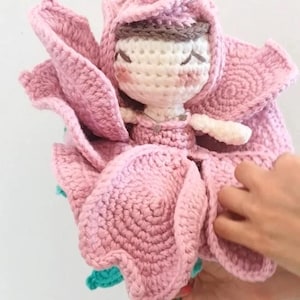 Può includere: Una bambola all'uncinetto fatta a mano a forma di fiore rosa. La bambola ha un viso bianco con gli occhi chiusi, guance rosee e una fascia marrone. Il fiore ha grandi petali a strati e uno stelo turchese.