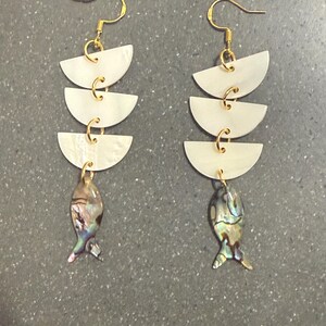 Peut inclure: Paire de boucles d'oreilles pendantes avec crochets dorés, trois éléments blancs semi-circulaires et un charm en forme de poisson. Le charm poisson présente des couleurs irisées bleues, vertes et violettes. Les boucles sont reliées par de petits anneaux dorés.