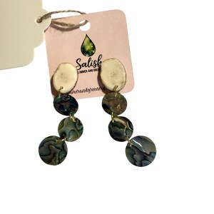 Peut inclure: Boucles d'oreilles pendantes avec une pierre supérieure naturelle blanc cassé et trois pierres circulaires irisées vertes et bleues. Les boucles sont reliées par des maillons en métal doré. Une étiquette rose indique "Salish Summer and Greenery".