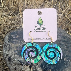 May include: A pair of circular earrings featuring an iridescent abalone shell inlay with a spiral cutout design. The earrings hang from gold-toned metal hooks and are attached to a light pink display card with the text Salish Glimmer and Greenery.