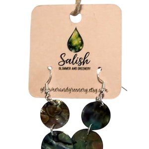Peut inclure: Boucles d'oreilles avec des pendentifs circulaires irisés dans des tons de vert, de bleu et de marron, suspendus à des crochets argentés. Les boucles d'oreilles sont présentées sur une carte beige avec le texte "Salish Glimmer and Greenery".