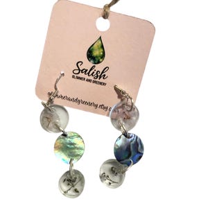 Peut inclure: Boucles d'oreilles pendantes argentées composées de cercles transparents, irisés et en nacre. Elles sont suspendues à une étiquette rose avec le texte "Salish Glimmer and Greenery" et l'adresse du site web.