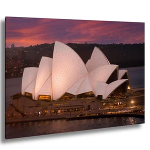 May include: The Sydney Opera House features white, sail-like roof structures illuminated against a dark purple and orange sunset sky. The building sits on the water's edge with visible architectural details and warm interior lighting at the base.