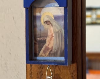 May include: A small, blue wooden frame with a religious image. The frame has a crown-like top with a blue and iridescent glass bead. A small, clear crystal hangs from the bottom. The image depicts a person with a halo.