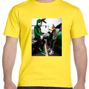 May include: A yellow t-shirt with a graphic print of Bugs Bunny and Daffy Duck wearing green hoodies and gold chains, standing next to each other in front of cars.