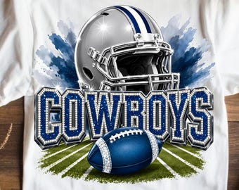 May include: A white t-shirt featuring a graphic print of a silver football helmet positioned above the word COWBOYS in large blue block letters with a white border. A blue football sits in front of the text on a green field graphic background.