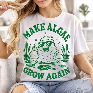 May include: A white short-sleeve t-shirt featuring a green graphic of a cartoon algae character wearing sunglasses, giving a thumbs up, surrounded by cattails and lily pads, with the text "MAKE ALGAE GROW AGAIN" printed in green.