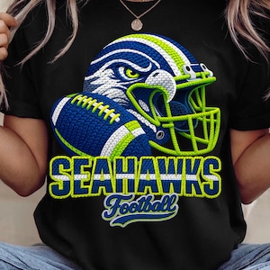 May include: Black t-shirt featuring a football helmet and football in blue, green, and white. The design includes the text "SEAHAWKS Football" in a bold, outlined font. The helmet has a detailed eagle design.