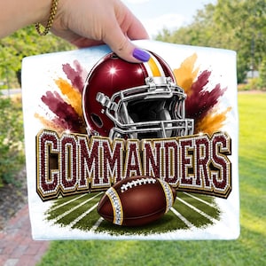 May include: A white t-shirt featuring a graphic of a burgundy football helmet with a yellow stripe, positioned above the word COMMANDERS in large, glittery burgundy letters. A brown football sits below the text on a green field graphic.