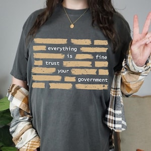 May include: Dark grey t-shirt with the text "everything is fine, trust your government" in a brick-like design. The shirt is worn with a plaid shirt and a gold necklace.