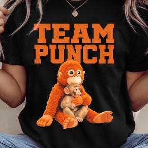 May include: Black t-shirt with the orange text "TEAM PUNCH" above a cartoon orangutan hugging a baby monkey. The orangutan is orange, and the baby monkey is tan. The shirt is a crew neck.