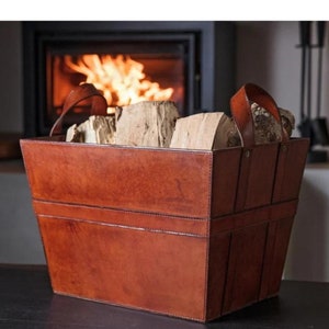 Puede incluir: Una cesta de leña rectangular de cuero marrón con dos asas, llena de leña. La cesta tiene un diseño cosido y está colocada frente a una chimenea con fuego.