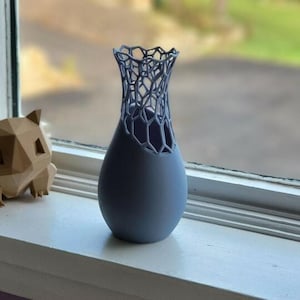 May include: A light blue vase with a unique openwork design at the top, sitting on a windowsill. A brown geometric figure and a purple musical instrument are also on the windowsill. The vase is egg-shaped.