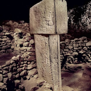Puede incluir: Un pilar de piedra tallada y desgastada con intrincadas tallas en relieve. El pilar es rectangular con una parte superior grande y plana. Las tallas representan figuras y patrones estilizados. La estructura se encuentra entre muros de piedra en bruto y un fondo oscuro.