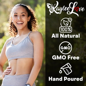 May include: A woman wearing a grey sports bra and leggings stands in front of a green background. The text "Kaylee Love" is written in white with a red heart. The text "100% All Natural" is written in white. A white circle with a red slash through it and the text "GMO" is written in white. The text "GMO Free" is written in white. A white outline of a measuring cup with a drop of liquid and the text "Hand Poured" is written in white.
