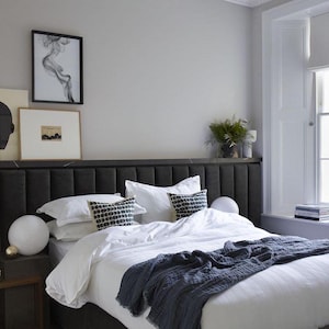 Könnte beinhalten: Ein weißes Bett mit einem dunkelgrauen Kopfteil und einer blauen Tagesdecke. Das Bett ist mit weißen Laken und Kissen bezogen. Neben dem Bett steht eine weiße Lampe auf dem Nachttisch.