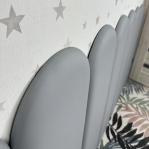 May include: A series of smooth, rounded, grey upholstered wall panels arranged in a vertical row. The panels feature a solid matte finish and are positioned against a white wall covered in a repeating pattern of light grey and beige stars.