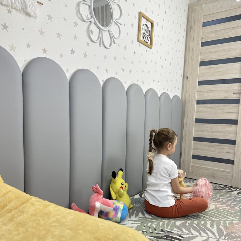 May include: A series of vertical, arched, light gray padded wall panels arranged side-by-side to form a continuous headboard-like structure. The panels have a smooth, matte finish and rounded tops, creating a soft, uniform, and clean architectural look.