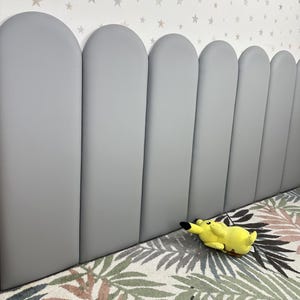 May include: A series of vertical, light gray, padded wall panels with rounded tops arranged in a row. A small, yellow, plush Pikachu character toy is positioned on the floor in front of the panels, which are set against a white wall with stars.