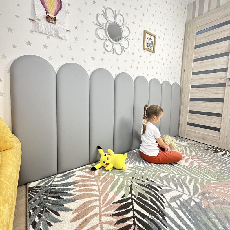 May include: A series of vertical, light gray, upholstered wall panels with rounded tops arranged in a row. The panels feature a smooth, matte finish and are positioned horizontally along the wall to create a continuous, padded headboard-like structure.